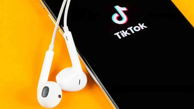 Photo of a phone with TikTok open, resting on a yellow background. A pair of white earphones are lying across it. Photo credit: Shutterstock