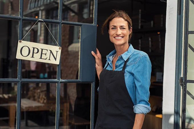 A light-skinned woman in a blue jean button-up and black apron holding a dark blue glass door open. The door has a tan sign on it that reads, "Open."