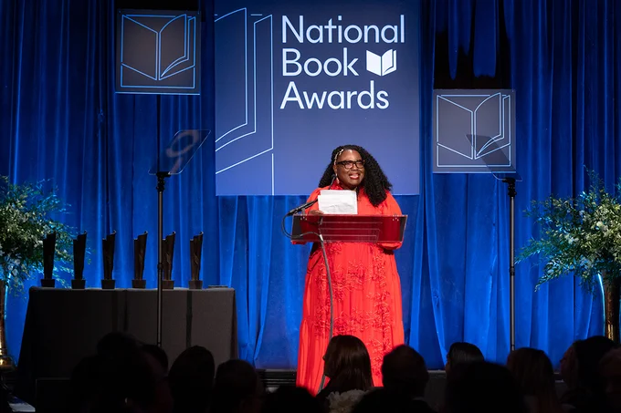 ALA Executive Director Tracie D,. Hall on stage at the National Book Awards Ceremony accepting the Literarian Award. Hall wears a bright red floor-length dress.