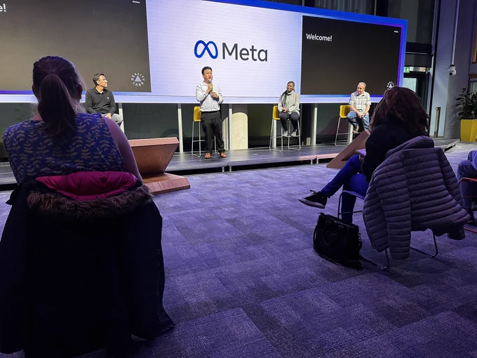 Alex stands on stage holding a microphone with three other speakers.
Behind them is the Meta logo