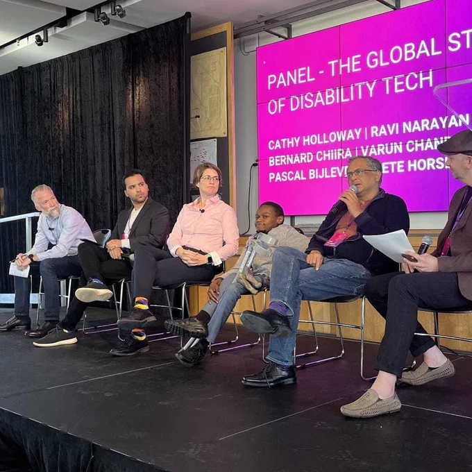 A panel of diverse people on stage with a screen behind displaying a purple slide with text “panel the global state of disability tech”