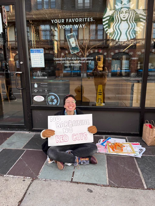 A smiling barista sits outside the Starbucks location at Coolidge Corner holding a sign that says “No Bargaining No Red Cups.” Next to them are a bunch of flyers and posters.