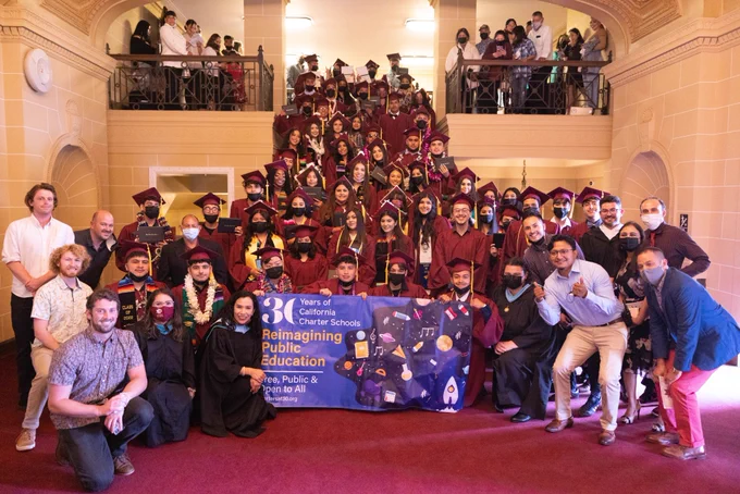 A large group of scholars in maroon graduate gowns with their supportive staff on the side and the charters 30 banner in the middle. 