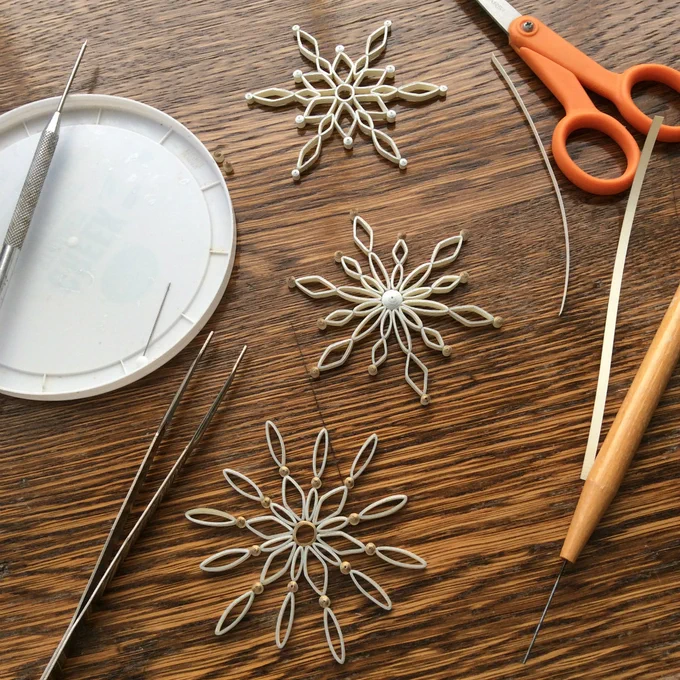 three silver, gold, and ivory quilled snowflake ornaments with quilling tools and paper strips on work table