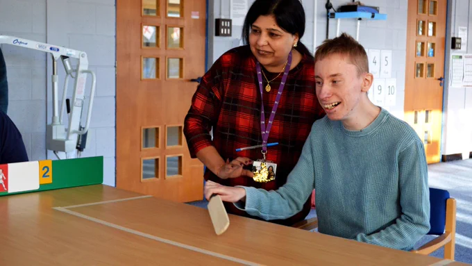 A young man smiles widely as he sits at a table holding a miniature cricket bat. On the table in front of him, a rectangle has been marked out in white lines. A support worker leans over his shoulder as they both wait eagerly for a ball to be played to him.