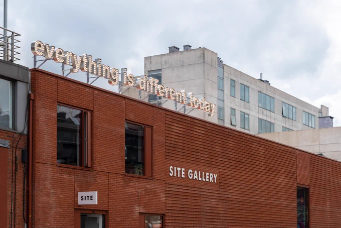 A red brick building with a sign on the front saying ‘Site Gallery’. On the roof, there is a metal sign with the words ‘everything is different today’ – each letter contains illuminated light bulbs.