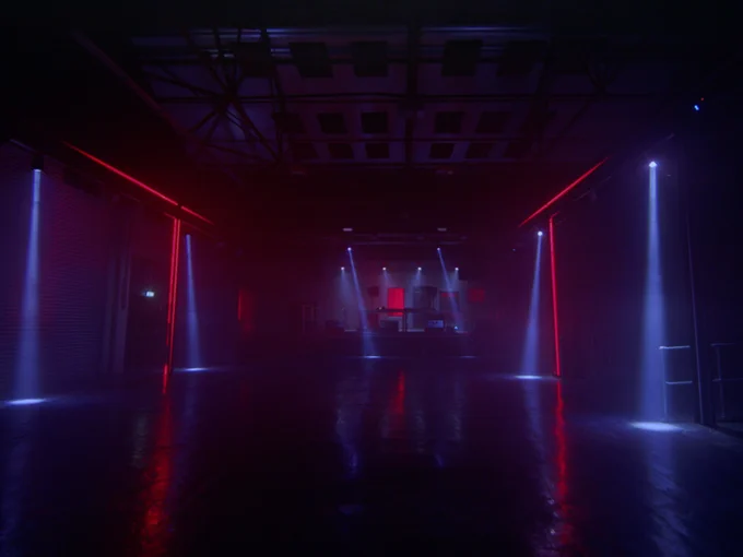 A dimly lit large dancefloor, looking forward towards a stage. Lines of red light are on the walls at each side, and beams of blue light from the ceiling hit the floor.