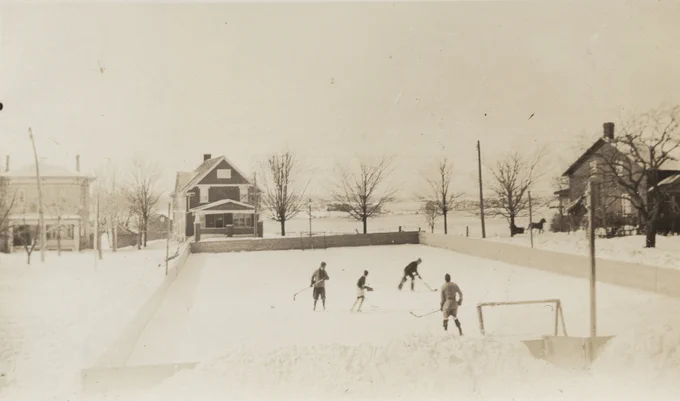 Four people playing hockey on an ice rink at the Evangelical Lutheran Seminary of Canada in Waterloo, Ontario. Albert Street can be seen in the background with fields beyond.