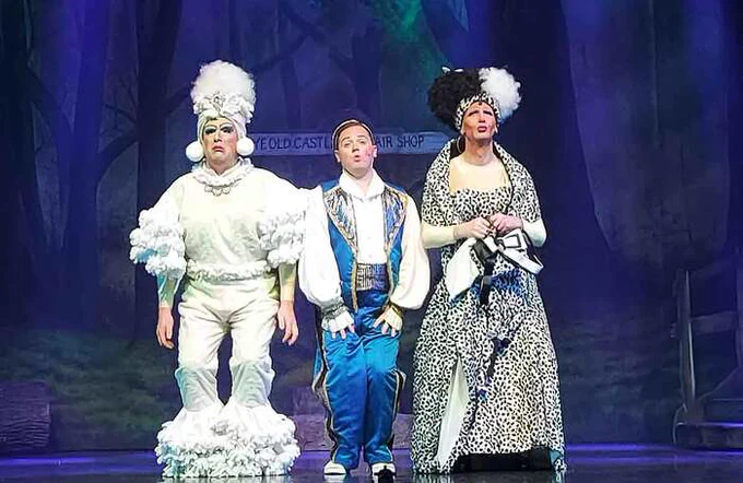 A scene from Cinderella at the Motherwell Theatre. There are three performers on stage in extravagant costumes, standing shoulder to shoulder singing to the audience