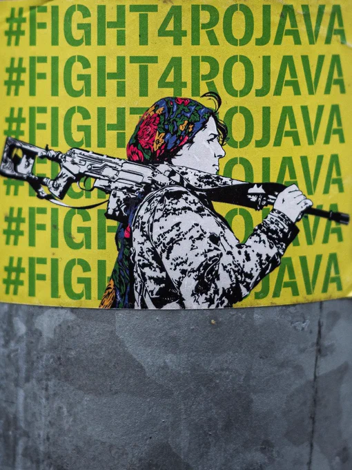 Yellow sticker on a pole taking over the whole photo. In the front a waist up monochrome drawing of a person which looks like a female kurdish fighter in combatsuit with a rifle on the shoulder and a colourful bandana on the head. The background reads six rows with #FIGHT4ROJAVA in green letters of which four rows are partially covered by the fighter.