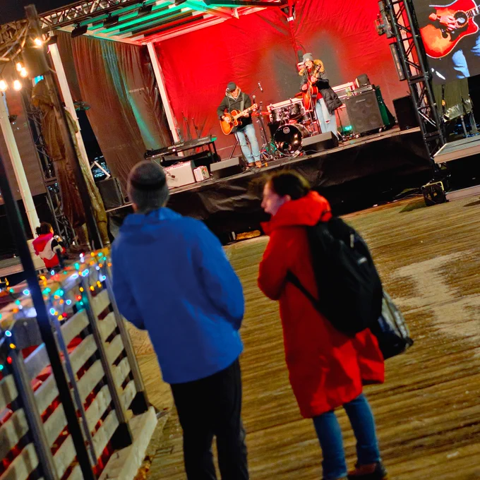 Two people stand on a wharf watching performers on stage.