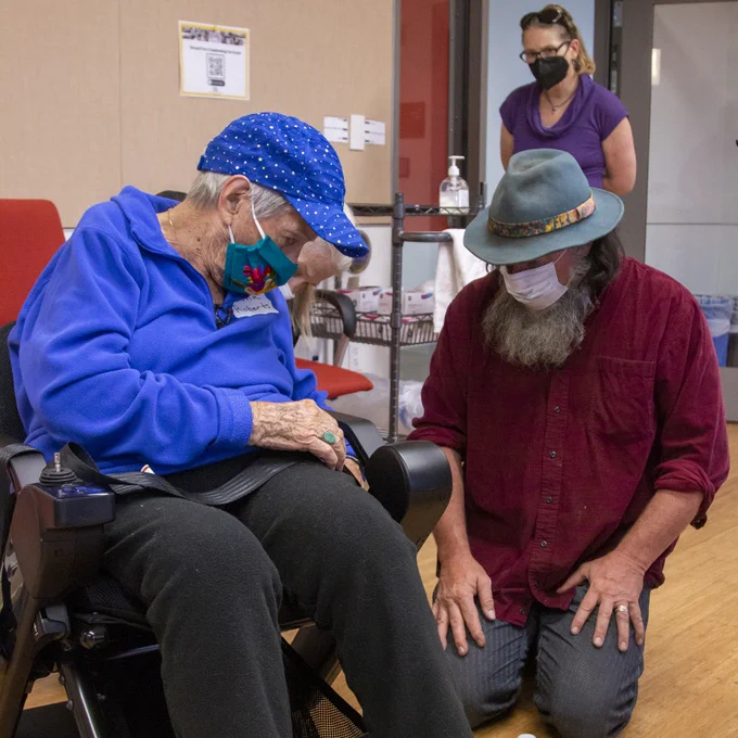 Photo of CIL Staff member John Benson examining Zona Roberts' wheelchair. John Benson is wearing a dark red button-up shirt, and Zona Roberts is wearing a blue sweatshirt.