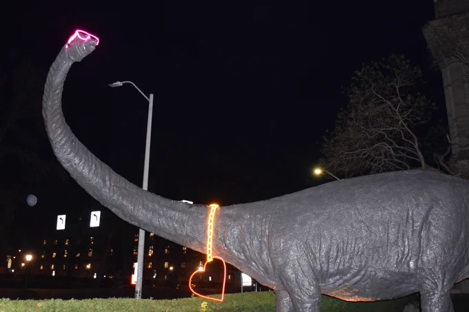 Dinosaur statue wearing pink neon shades and a light-up red heart necklace. 