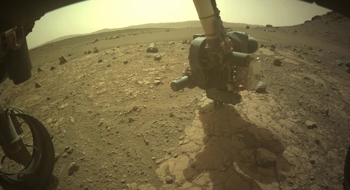 Photo from wide-angle camera mounted low on the front of NASA’s Mars Perseverance rover shows the rover’s robotic arm holding its coring drill against a flat patch of rock on Mars. Also visible in the bottom left corner of the image are parts of the rover’s left wheels.