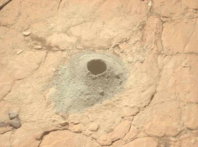Close-up photo from NASA's Mars Perseverance rover shows a flat patch rock on Mars, with a drill hole where it has collected a rock core sample. A small cone of powdered drill tailings surrounds the hole.