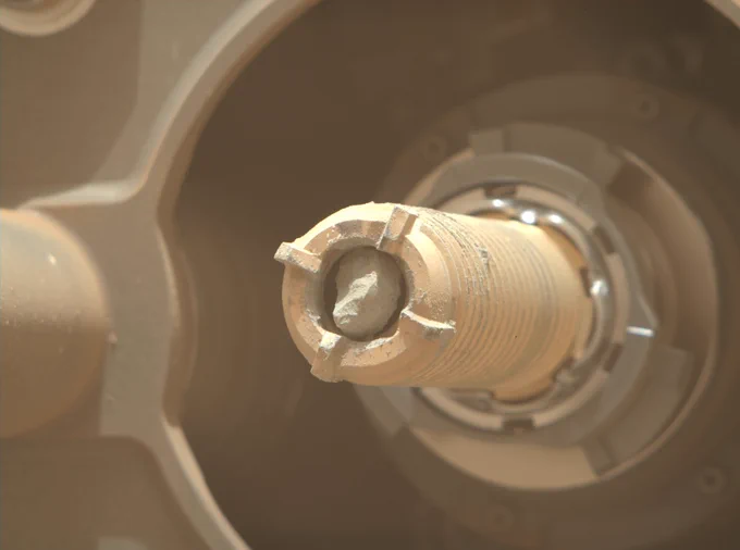 Close-up photo from NASA’s Mars Perseverance rover looks at the rover’s drill bit from an oblique angle. A sample of rock core sits inside the hollow, gold-colored bit, which has teeth around its front edge, and a spiral pattern along it.