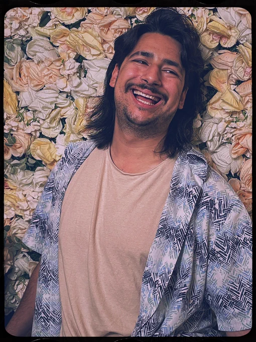 J. Chavez headshot, of a smiling person with dark mid length hair and moustache, wearing a peach tshirt under a blue patterned shirt, against a floral background.