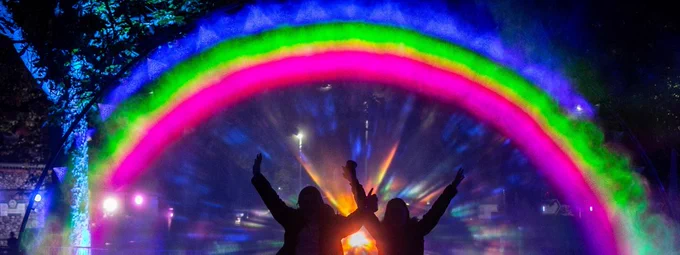Two people standing in front of a rainbow, their arms are raised in the air