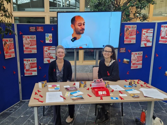 Abi and Caro in the atrium of the USB for World Aids Day. They have a table in front of them with ribbons and post cards, and behind them a video from the project. And some posterboard with posters about World Aids Day. 