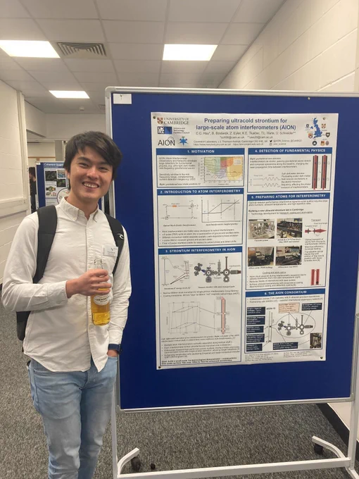 A person stands smiling and holding a bottle of cider in front of a poster board with a poster entitled 'Preparing ultracold strontium for large-scale atom interferometers (AION)'. They are in a narrow poster hall at a conference and another poster is visible in the background. 