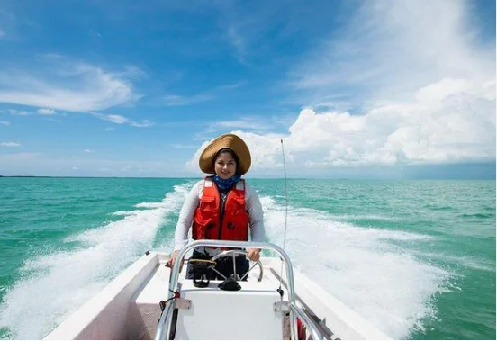 A person in a large hat, wearing an orange lifejacket vest seers a boat. There is teal, open water behind the boat. The boat is creating a white splash of water behind it. The sky is blue with puffy white clouds. 