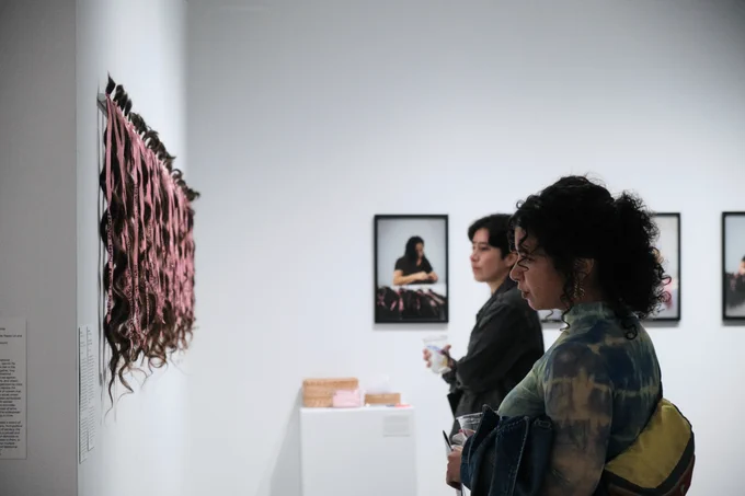 In a white gallery space, two people on the right side of the frame look hard at an installation on the wall on the left. Many strands of hair with pink ties are hung vertically. Photographs are hung in the background.
