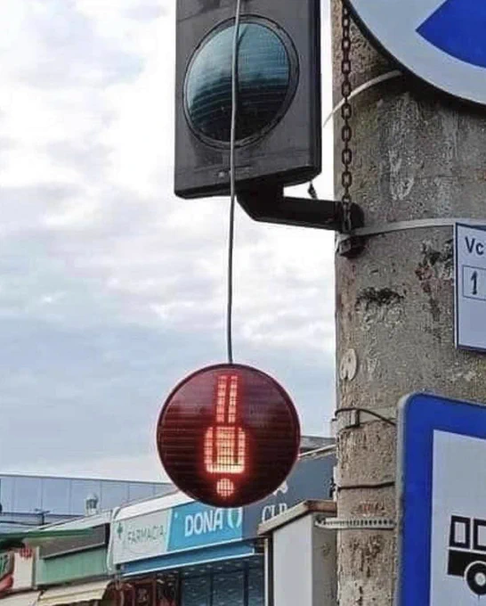 A traffic light showing the pedestrian lights, the light showing the red standing person has fallen out of its socket and is hanging upside down (but still lit) below the trafic light. 