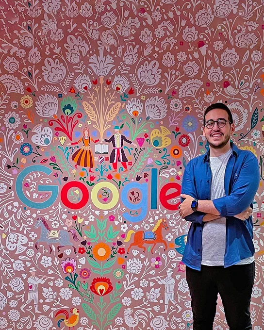 Rand wearing glasses in a grey shirt and blue button up smiling in front of a multicolored Google sign.