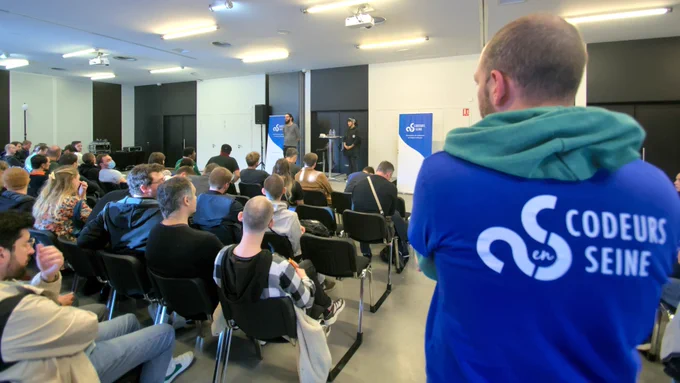 Plan d’ensemble avec au premier plan à gauche le public, à droite un bénévole dont on peut voir le tee-shirt avec le marquage « Codeurs en Seine ». En arrière-plan, Noël Macé et Valériane Venance sur la scène sont en train de répondre aux questions du public.
