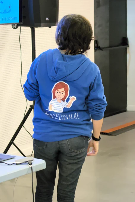 Aurélie Vache de dos avec un sweat à capuche sur lequel est dessiné un personnage à son effigie sous-titré avec son compte Twitter