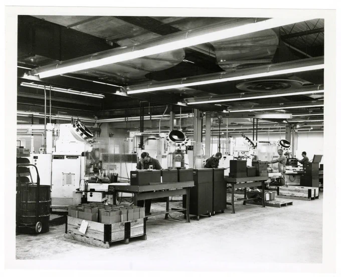 Five workers, tables, tools, and equipment at the machine shop of CDC in Minneapolis in 1968. The room is large and there is plenty of space for the workers to spread out and do their design and manufacturing work.