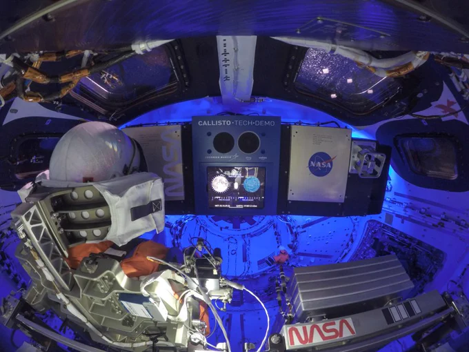 A view inside the Orion spacecraft shows many screens, wires, and the Callisto Tech Demo. The spacecraft is filled with blue light and Moonikin Campos, a manikin with a white helmet and orange suit.