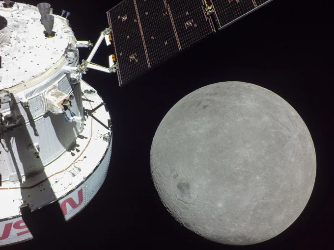 The Orion spacecraft captures an image of the Moon. The far side of the Moon looms large just beyond the spacecraft. The image showcases the black background of space, while the Moon shines grey on the right, and the white structure of the spacecraft is shown on the left. 