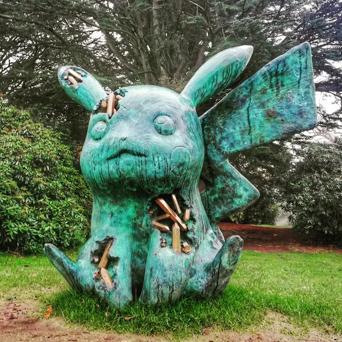 Bronze sculpture of Pikachu - green patina surface with holes revealing gold rod like crystals. Sat in front of green bushes and trees in gardens. 