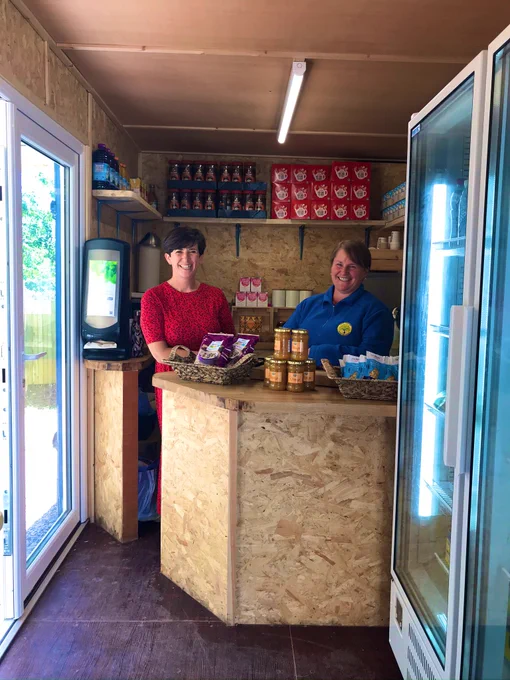 Two smiling volunteers stand behind the counter in the shop. The walls and counter are wooden and on the shelves are teas and coffees. A massive fridge is in the foreground.