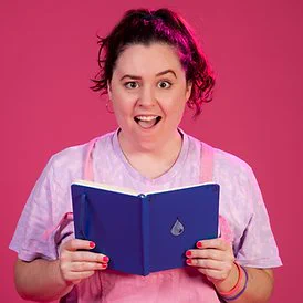 Promo photo for my show. I'm a white person with dark wavy hair in a high ponytail, in pink dungarees against a pink backdrop holding a blue book open with a teardrop on front. The expression on my face is mock-shock, open mouth, one eyebrow raised, as though scandalised by the contents of thr book (its my teenage diary) 