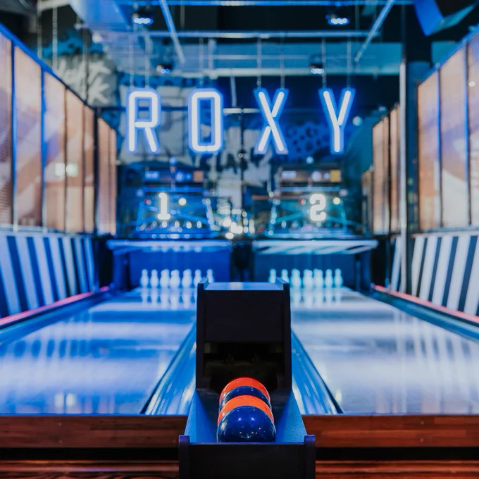 A neon lit bowling alley with two lanes and a ball dispenser in the middle. The word ‘ROXY’ hangs over the lanes in bright neon lighting.