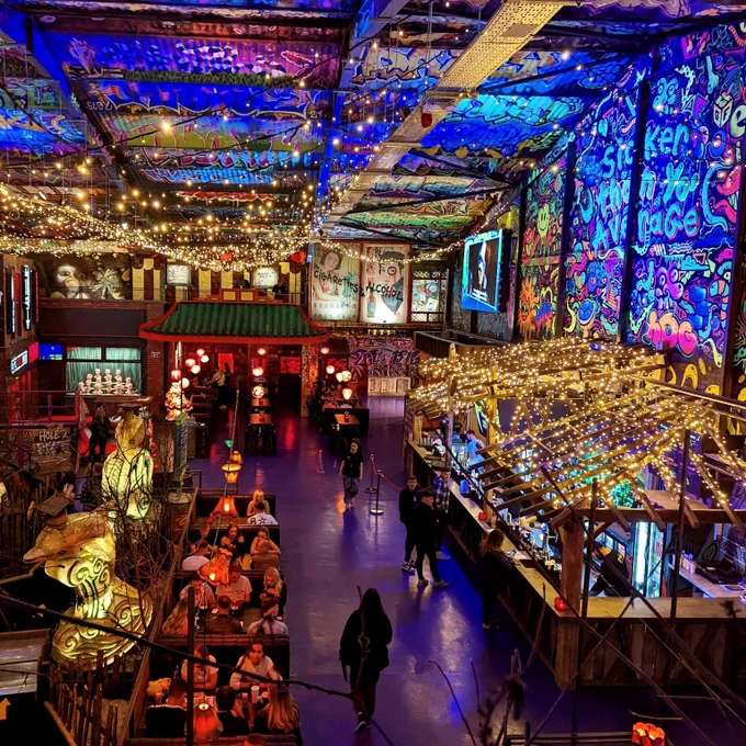 Golf Fang Sheffield - A large room with metal ceiling, lit by thousands of fairy lights. The walls and ceilings are covered in graffiti. A bar is on the right side, with seating booths in the centre. On the left side is one of the mini-golf courses.