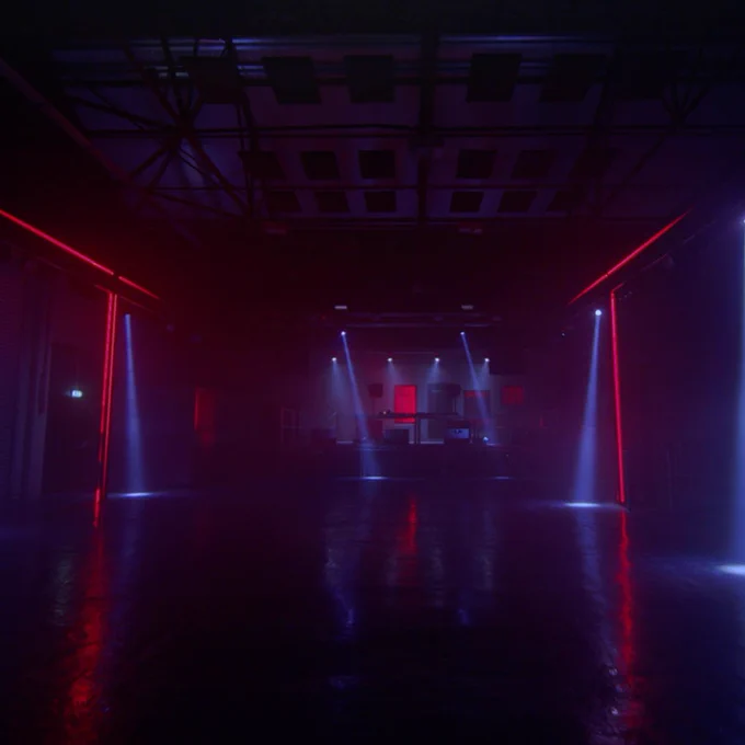 A dimly lit large dancefloor, looking forward towards a stage. Lines of red light are on the walls at each side, and beams of blue light from the ceiling hit the floor.