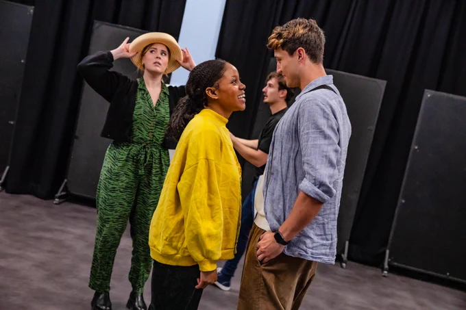 Three performers – Amara Okereke, Jordan Luke Gage and Ceili O'Connor, in a rehearsal room. Okereke is smiling at Gage while O'Connor holds her hat. 