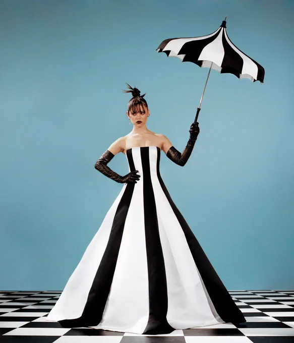 Dazzling photo of Jenna Ortega wearing a long black and white striped gown, black elbow length gloves, and a bold black lipstick while holding a black and white umbrella and standing on a black and white checkered floor. 