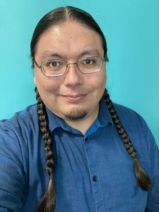 Alan has his long hair braided into 2 braids, wearing a blue shirt. He has a happy face with a little smile.