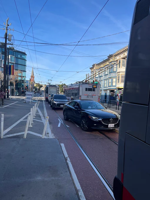 Photo of a transit only lane at church and market, with a 22 bus stopped at the platform and 3 personal vehicles behind it, followed by the J Church train forced to block some lanes on market street. 