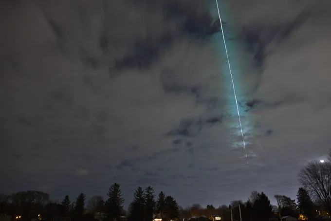 The sky at night is shown with a lot of cloud cover. Toward the bottom of the image are pine trees stretching out into the grayish-dark blue sky. On the center-right side of the image is a bright white line that has a teal-colored outline. This streak of light across the sky is a tiny asteroid that harmlessly entered Earth's atmosphere. To the very right of the image toward the bottom half, a partially full moon with a C-shape over the top half can be seen over a twiggy, bare tree.