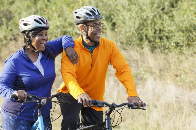 Two people smiling looking into the distance as they stand with their bikes in a park. The women is resting her arm on the man's shoulder.