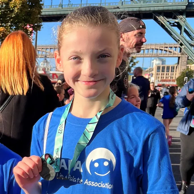  [Image ID: A photo of a girl wearing a blue CLAPA t-shirt holding a medal at the Great North Run] 