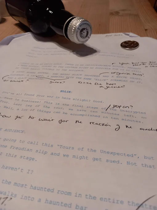 A picture of the annotated script for 'Unscrypted', a monologue by myself. A bottle sits sideways on the table nearby (to the left). To the right, on the top corner of the script, is a 10p piece. The name "Ellie" is clearly visible.