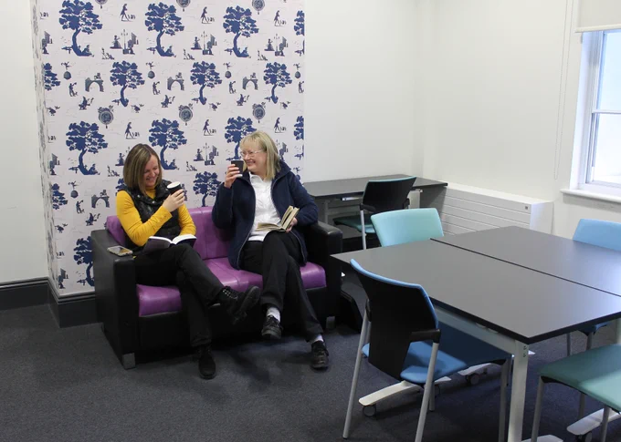 Two ladies are sitting on a purple sofa in a spacious room; they are smiling at each other, each holding a book and a cup with a hot drink. In front of them are tables with comfortable chairs. 