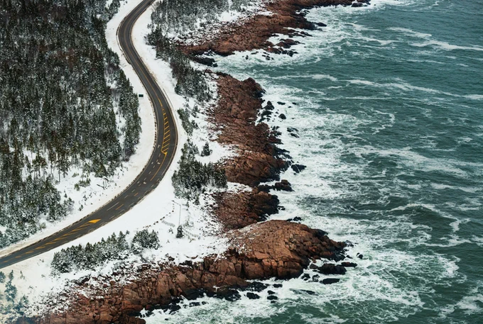 Waves wash against a rugged shoreline, beside a snow-covered roadside that sits opposite dense woods. 