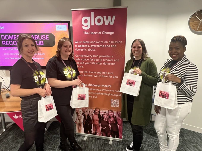 Glow staff members wearing Glow tops, standing by a Glow banner. They are holding white paper bags and smiling at the camera. 